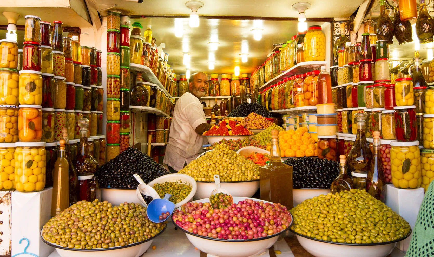 A Marrakech olive vendor surrounded by colorful pickled lemons, peppers, and towering bowls of olives.