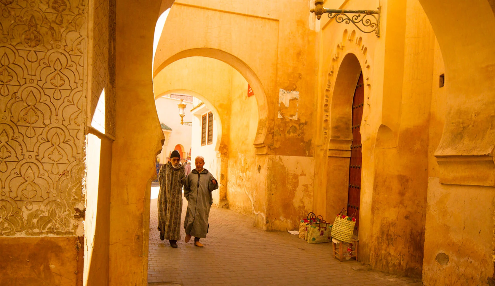 Two men walking beneath sunlit arches in Marrakech's medina near the Ben Youssef Madrasa.