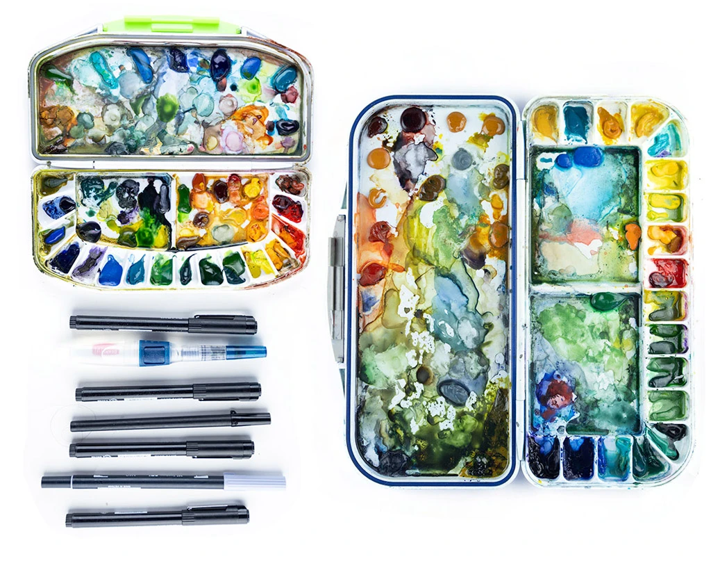 Two well-used travel watercolor palettes with mixed paint wells beside a row of black ink pens.