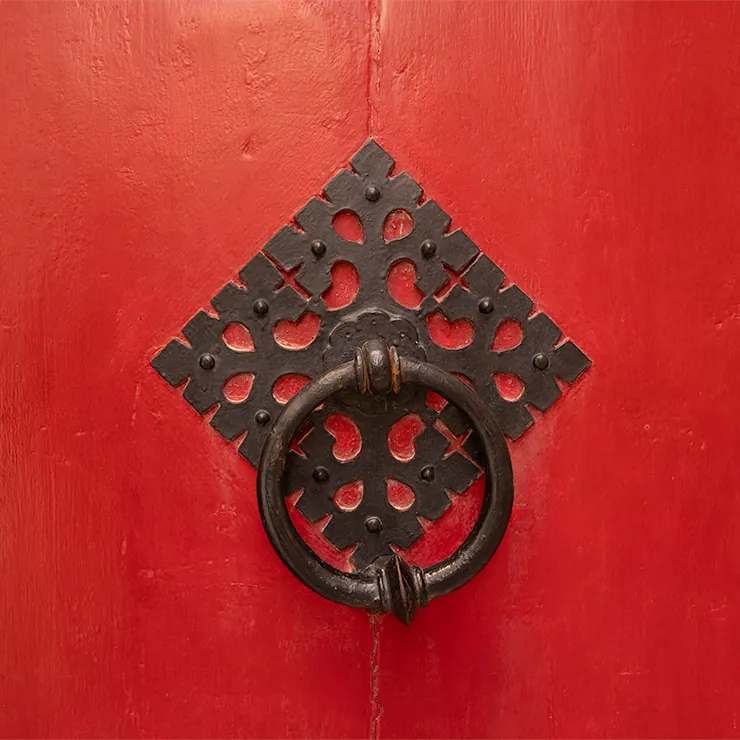 Ornate black iron ring door knocker on a bold red wooden door in Mdina, Malta, mounted on a decorative diamond-shaped iron backplate with intricate cutouts and small raised rivets.