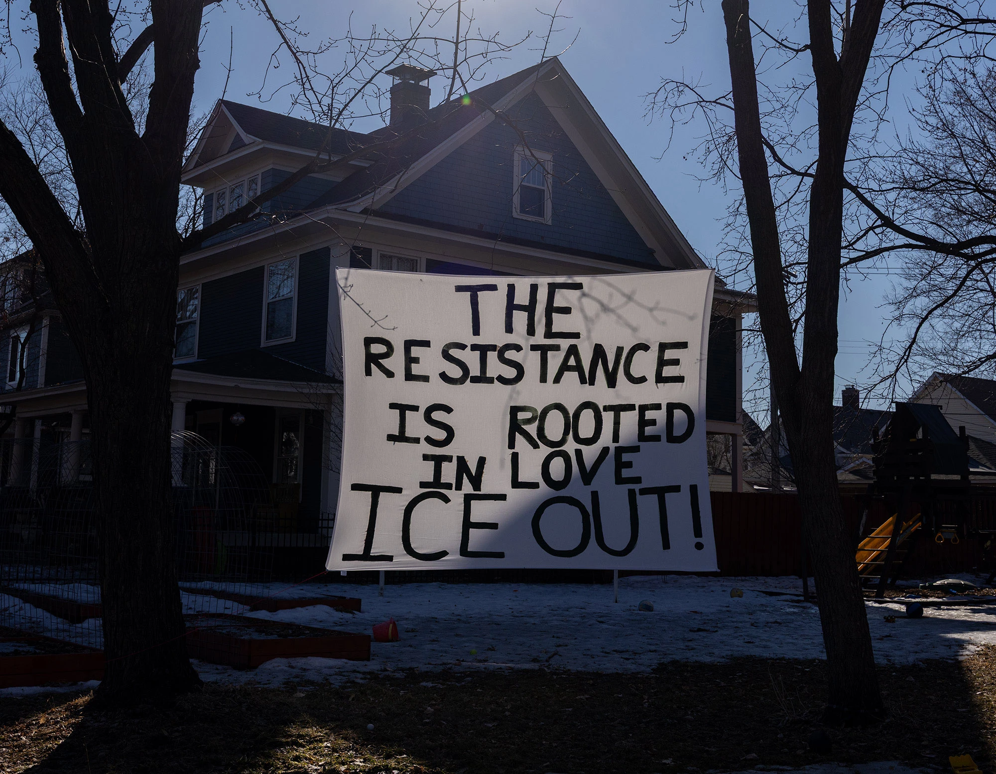 A large handmade sign strung between trees in a Minneapolis neighborhood reading 'THE RESISTANCE IS ROOTED IN LOVE ICE OUT!'