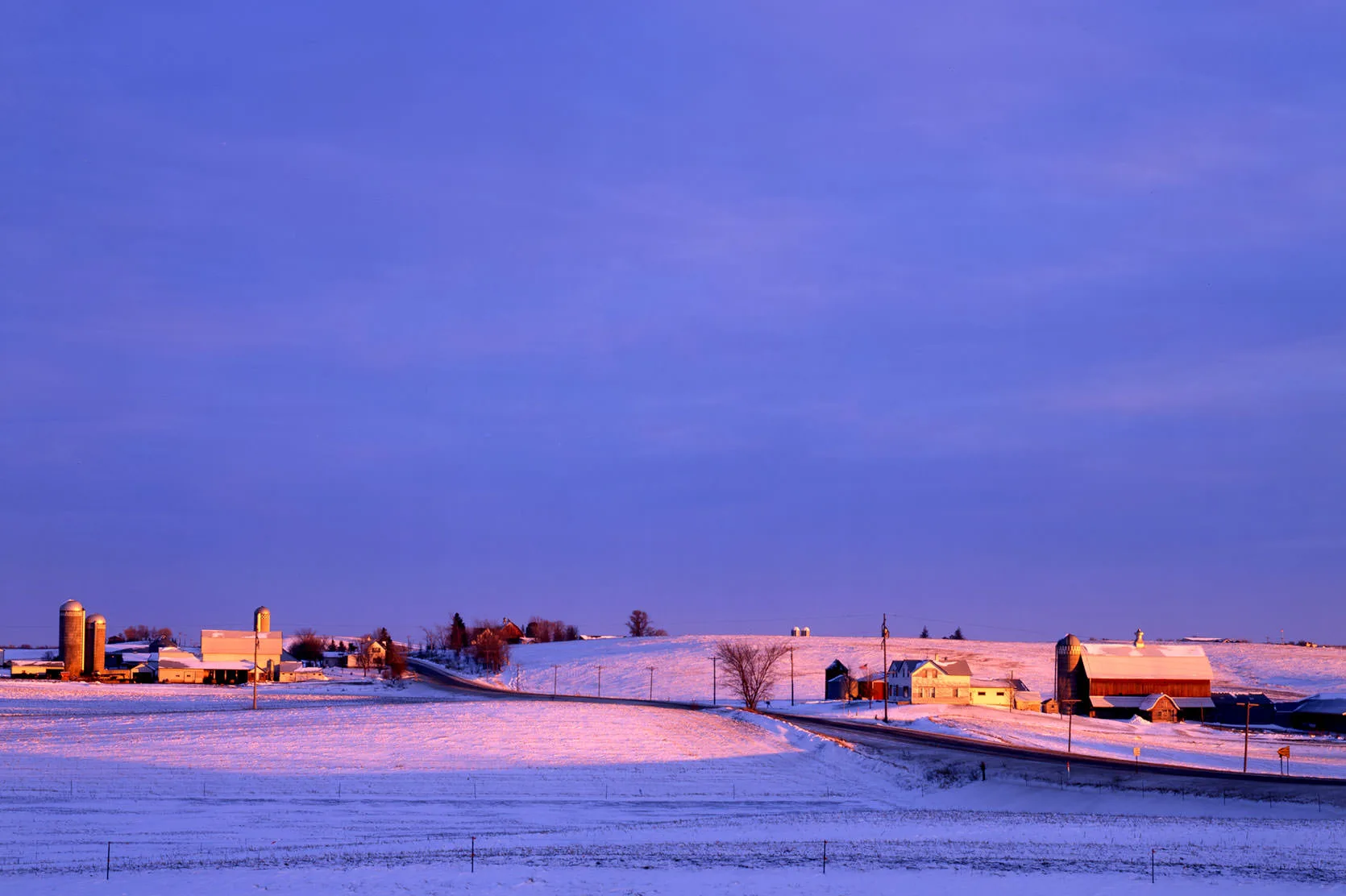 Red barns, silos and farmhouses glowing in low winter light across snowy Minnesota farm fields.