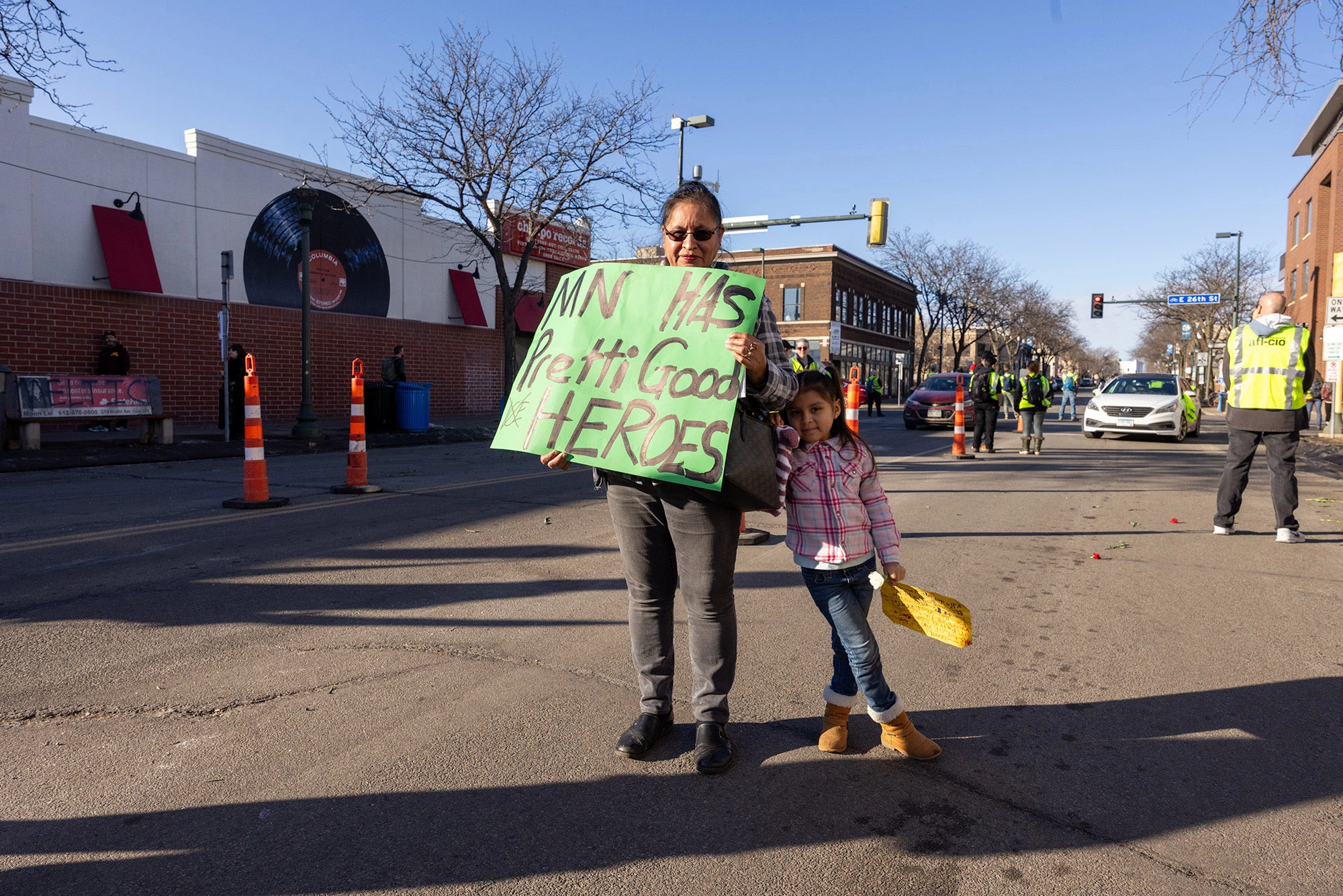 A Latina woman and her young daughter standing on a Minneapolis street holding a sign that reads 'MN has Pretti & Good heroes' during the Alex Pretti memorial.