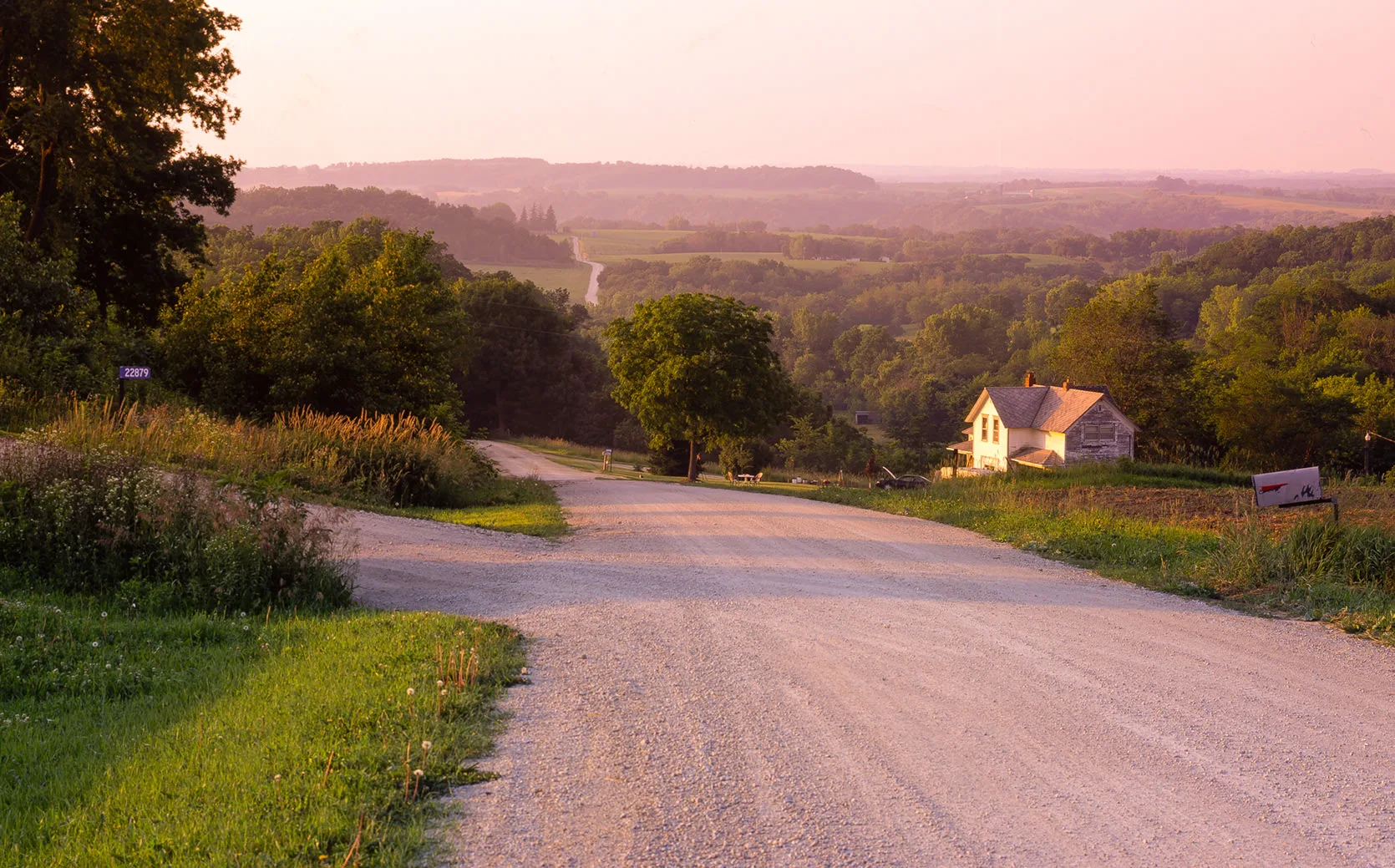A gravel road dropping through rolling green farm country at pink dusk, an old farmhouse below, southeastern Minnesota