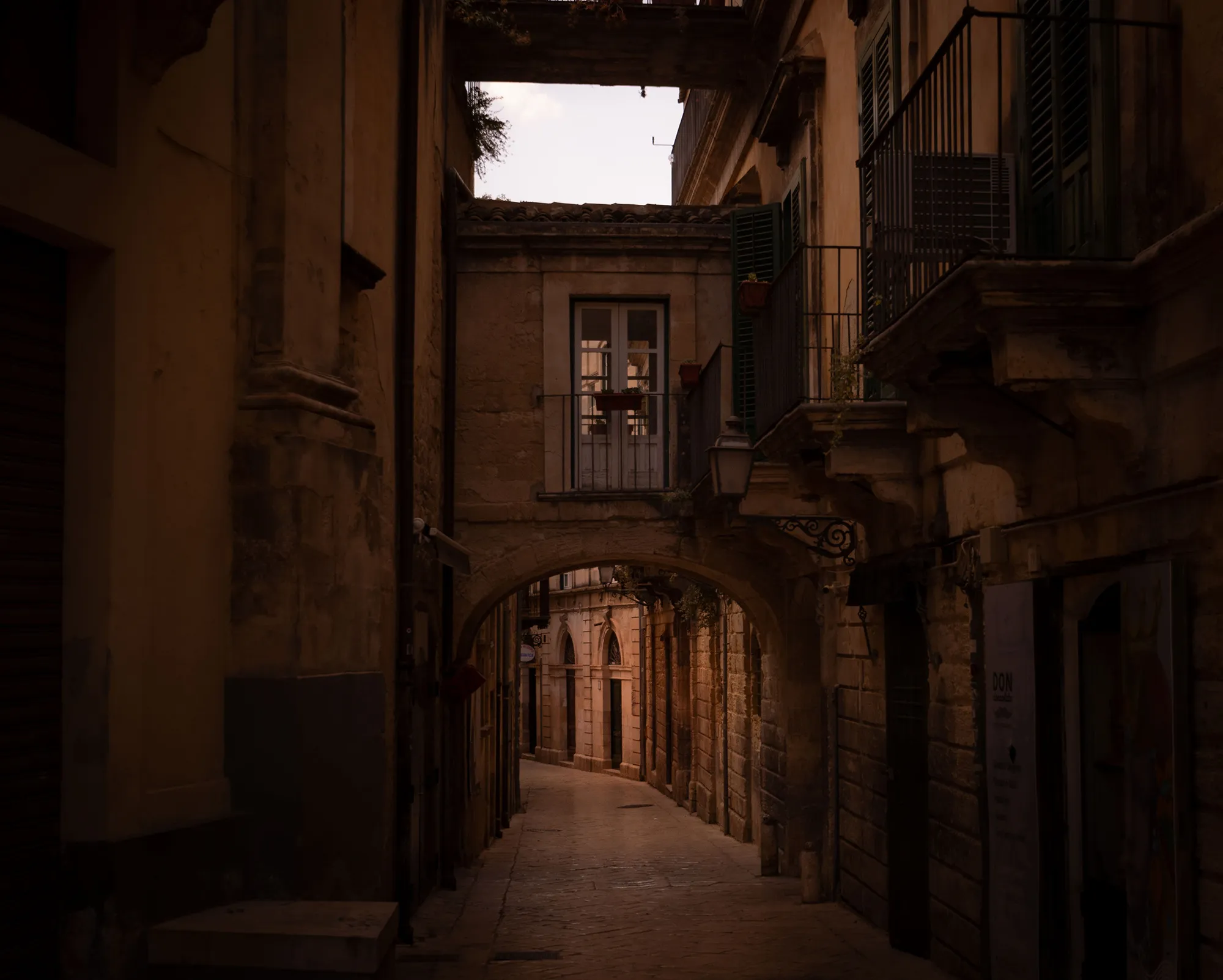 A stone arch beneath a covered passage in Modica, a narrow limestone lane running on into shadow past iron balconies.