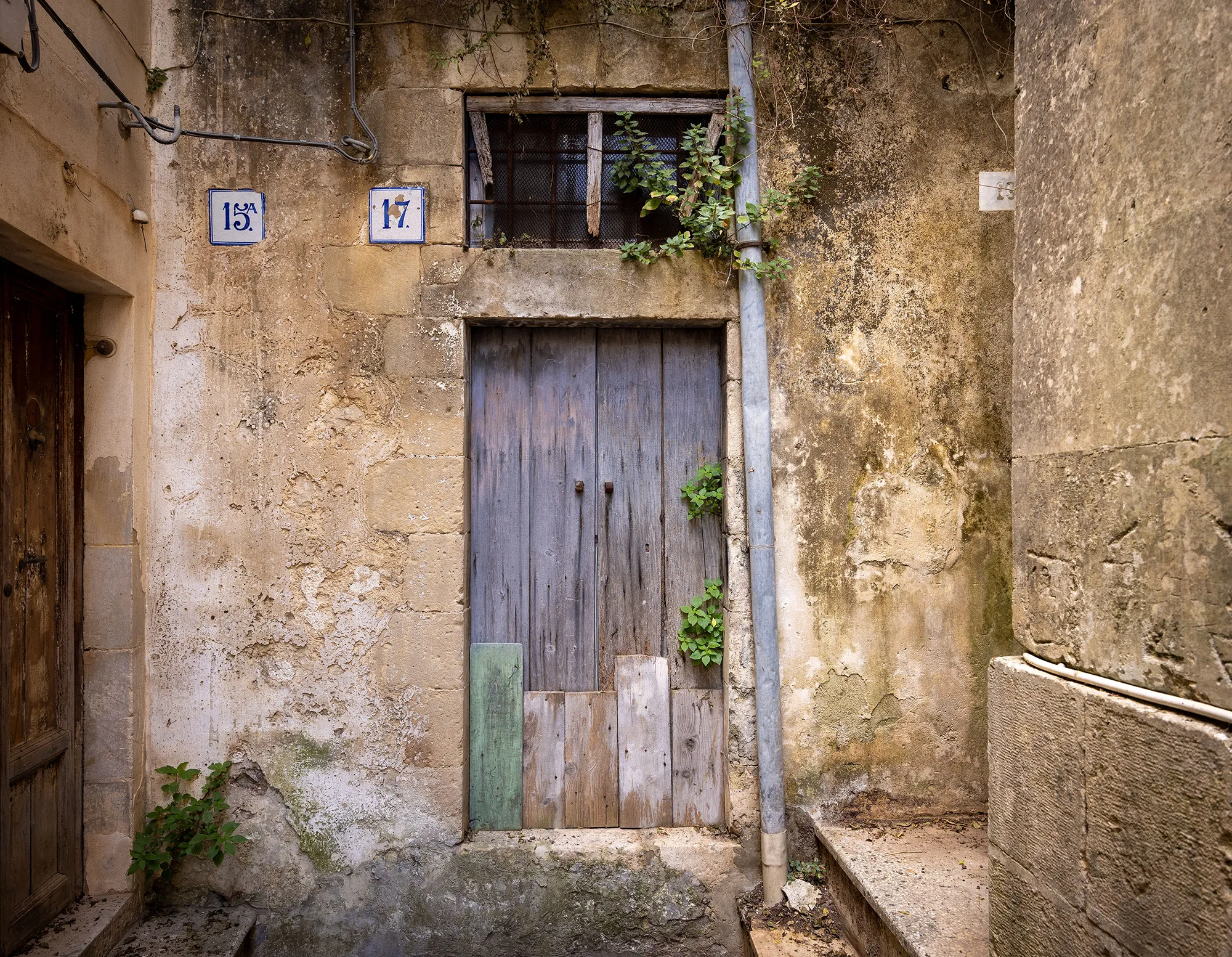 A weathered blue-gray wooden door patched with odd planks in a crumbling limestone wall in Modica Bassa, with blue-and-white house numbers and a barred window above.