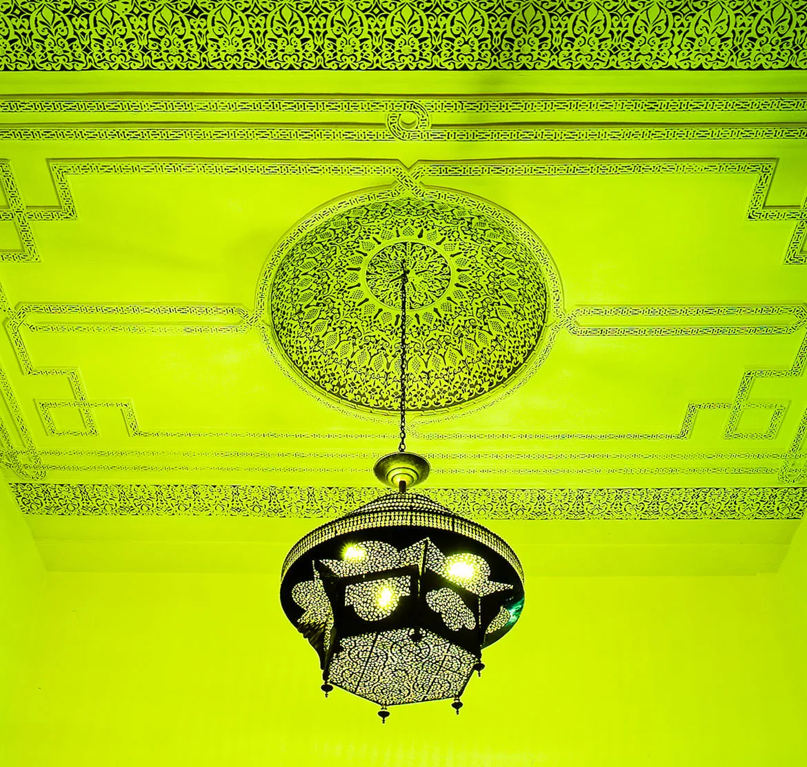 An ornate pierced-metal Moroccan lantern hanging from an intricately carved ceiling, lit vivid green