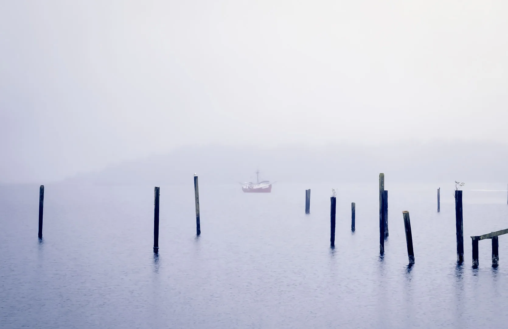 Old pilings and a lone fishing boat in dense fog on Neah Bay harbor.