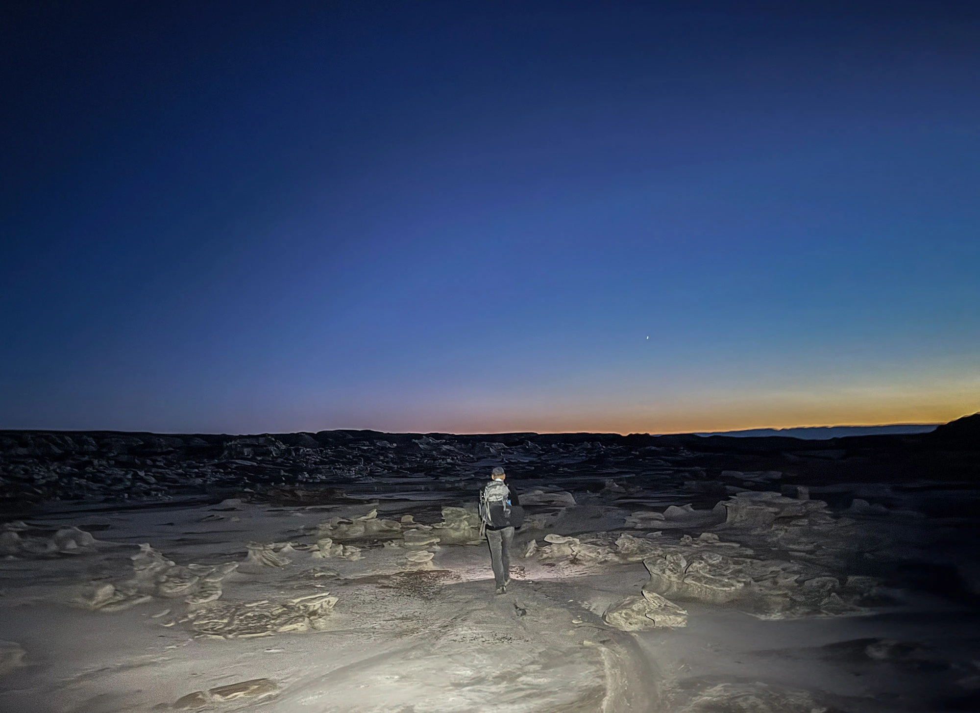 A hiker with a headlamp crossing the pale hoodoos of the Bisti Badlands at dusk, an orange band of afterglow on the horizon