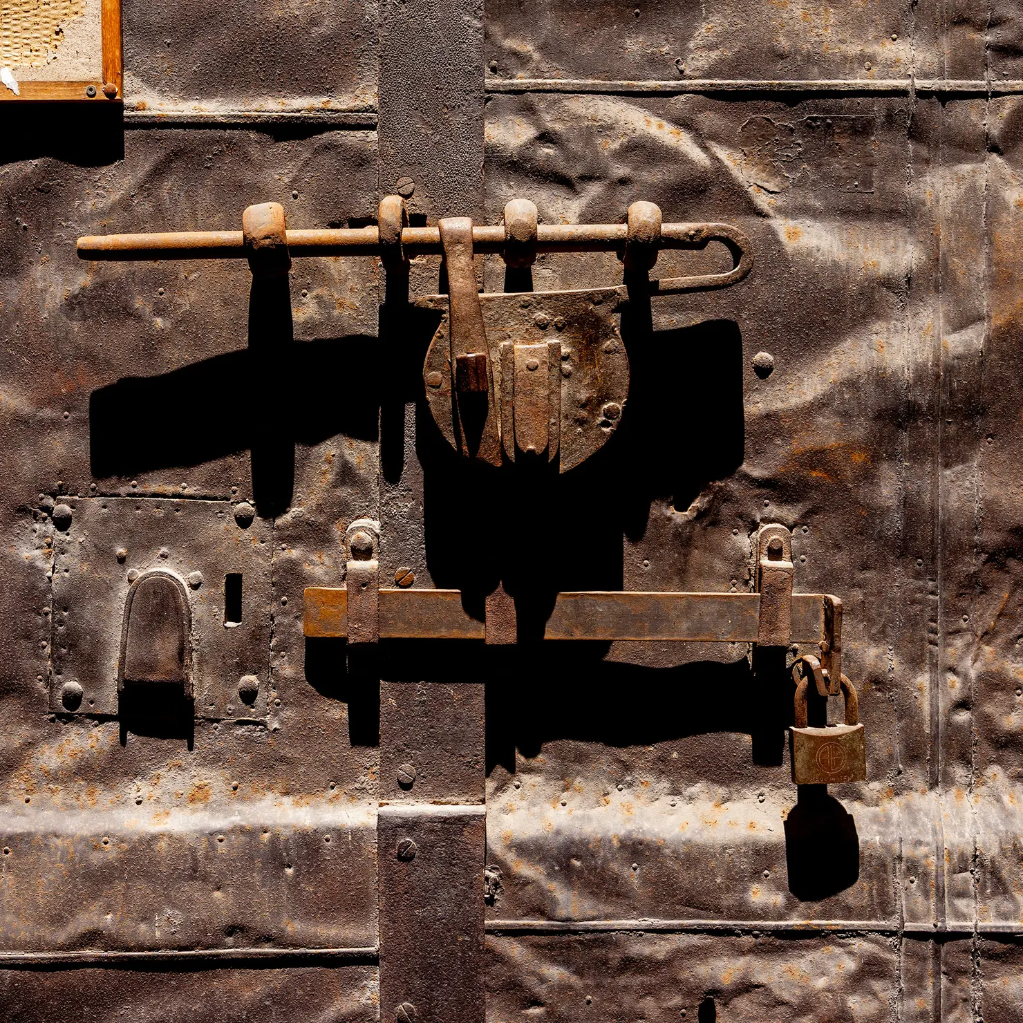 Iron bolts, hasps and a padlock layered on a dented sheet-metal door in Palermo, Sicily.