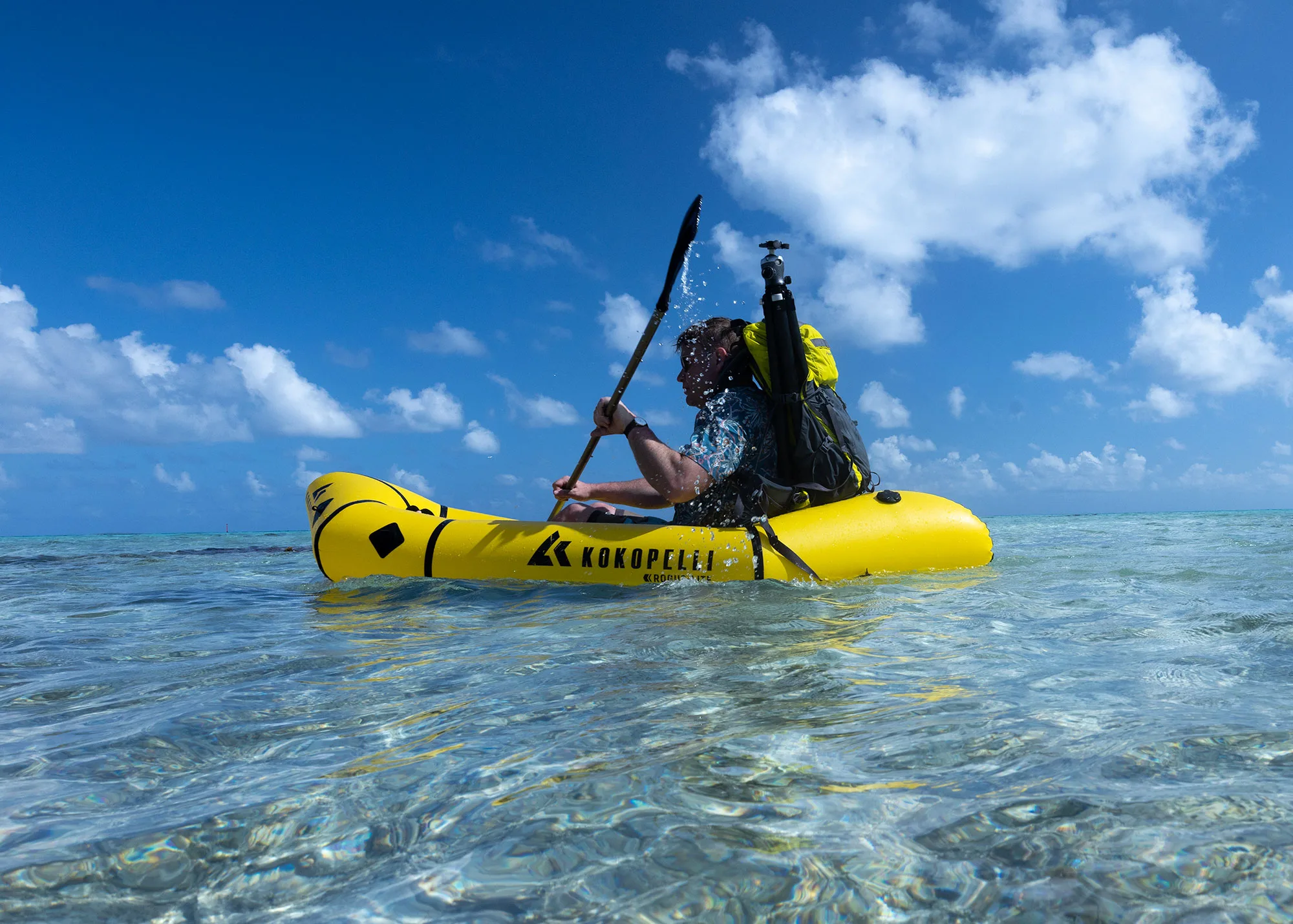 A paddler in a yellow Kokopelli packraft on clear turquoise water, holding up a camera in a waterproof housing.