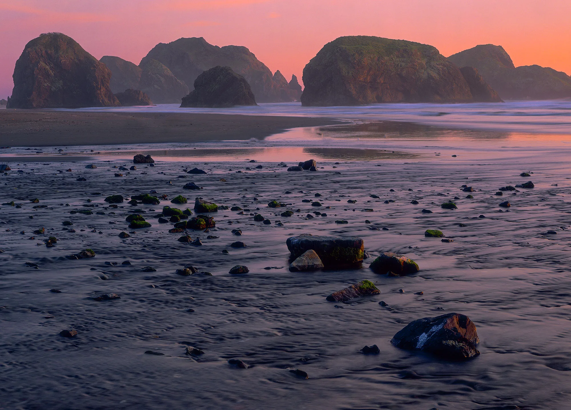 Sea stacks and scattered rocks at Pistol River beach on the southern Oregon Coast, under a pink sunset sky reflected on wet sand.