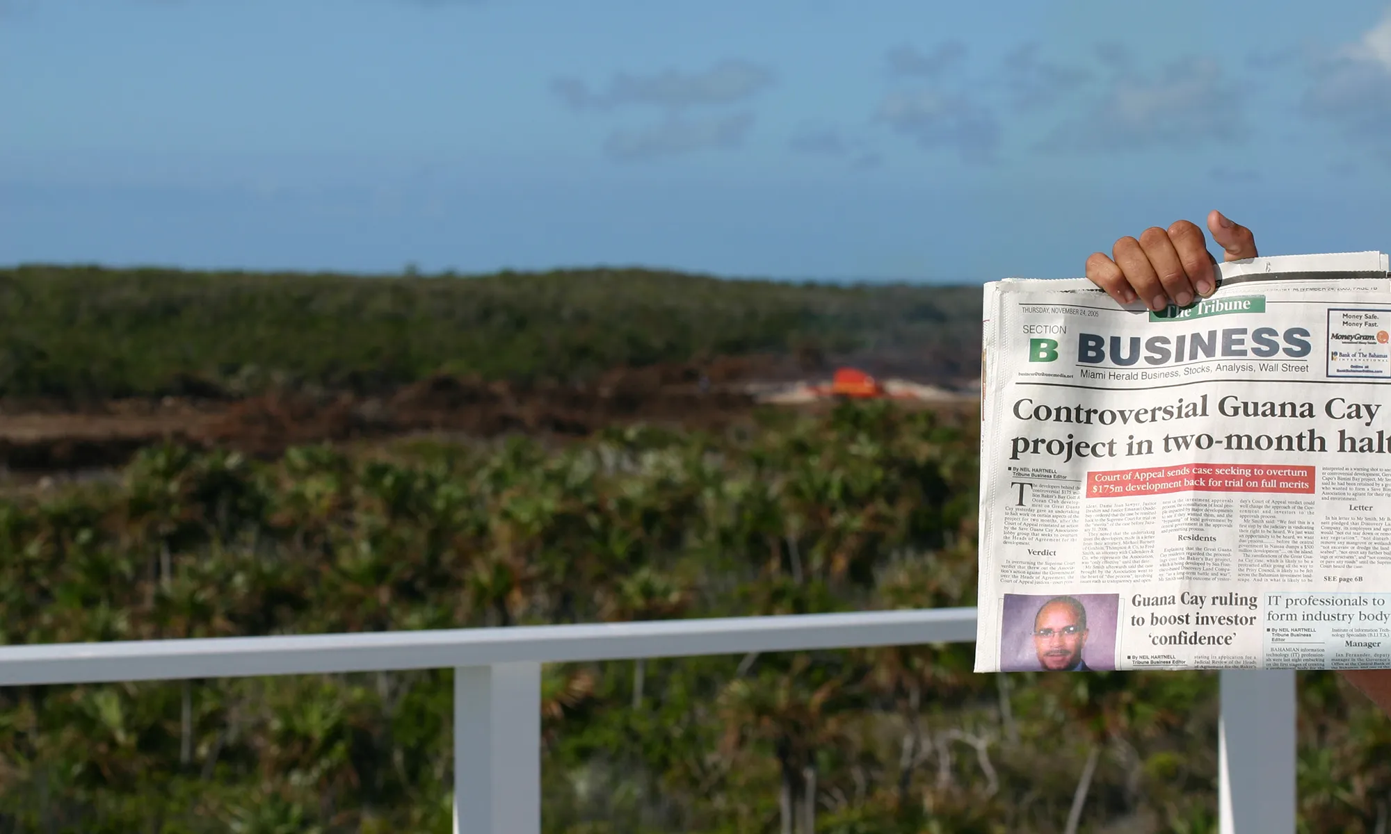A newspaper headline, “Controversial Guana Cay project in two-month halt,” held up in front of the cleared Bakers Bay site