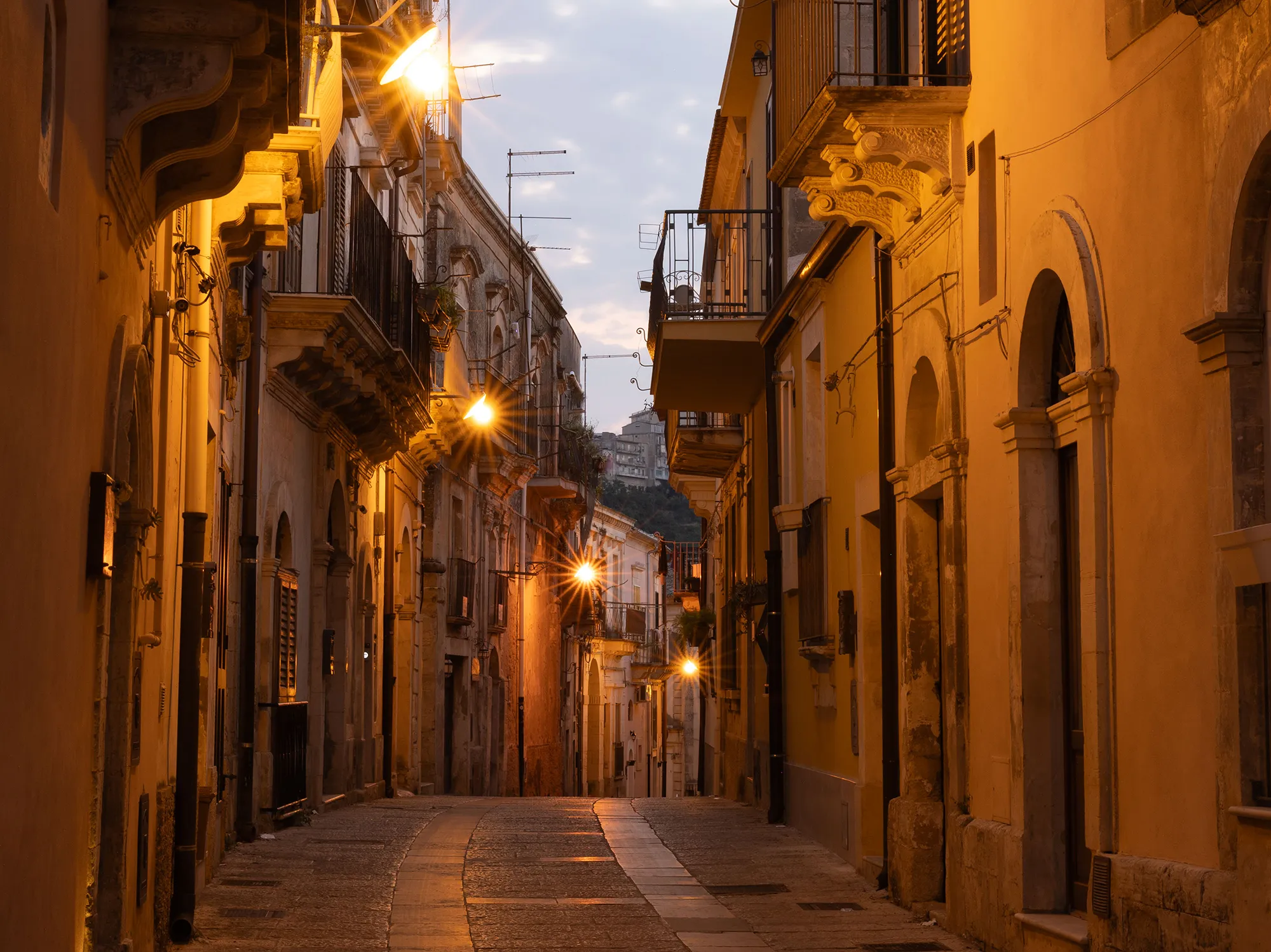 Streetlamps glowing along an empty limestone street in Ragusa Ibla at dusk.