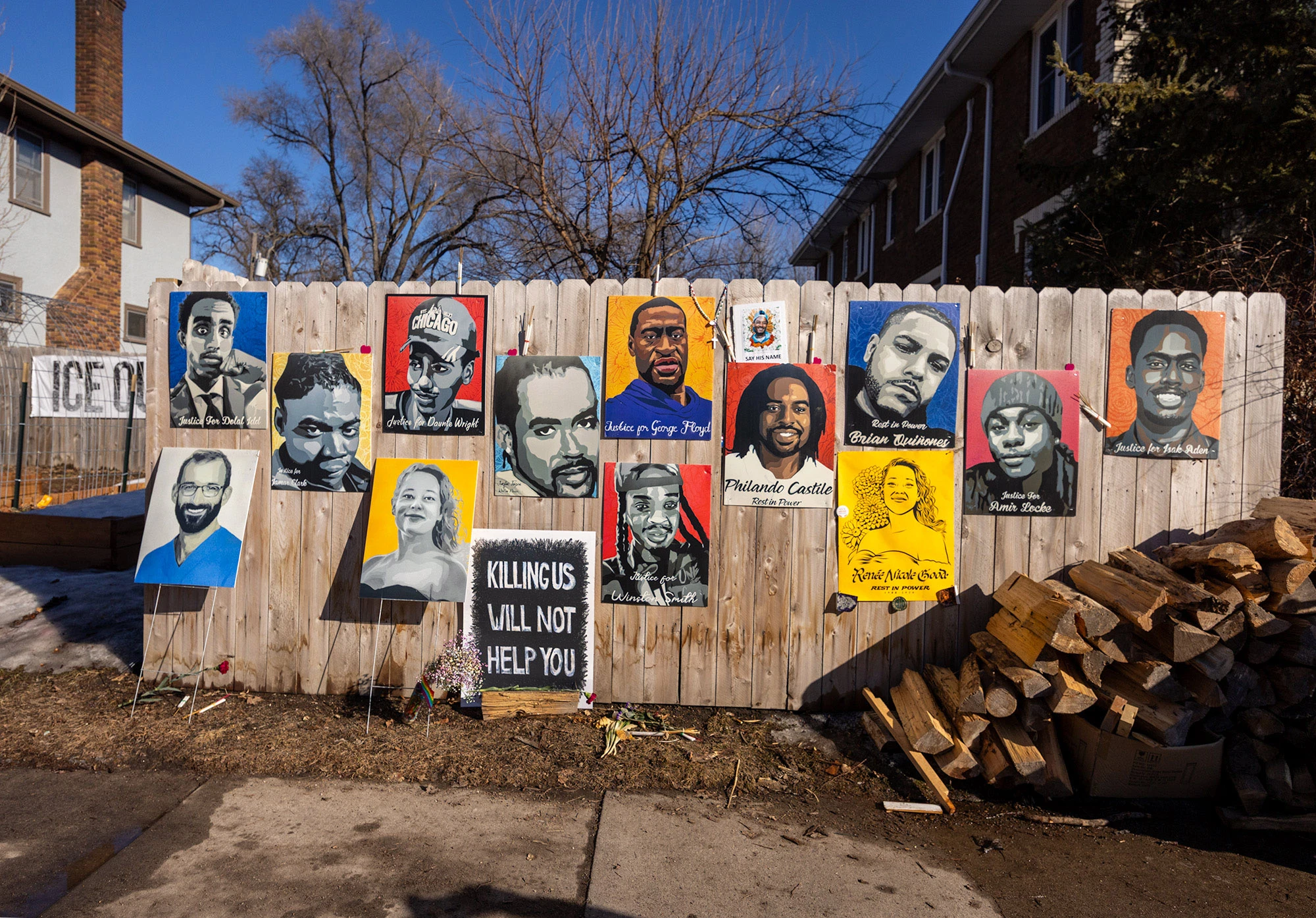 A fence outside the Renee Good memorial in Minneapolis hung with painted portraits of people killed by police and federal agents, including Renée Good, alongside a crowd gathered at the site.