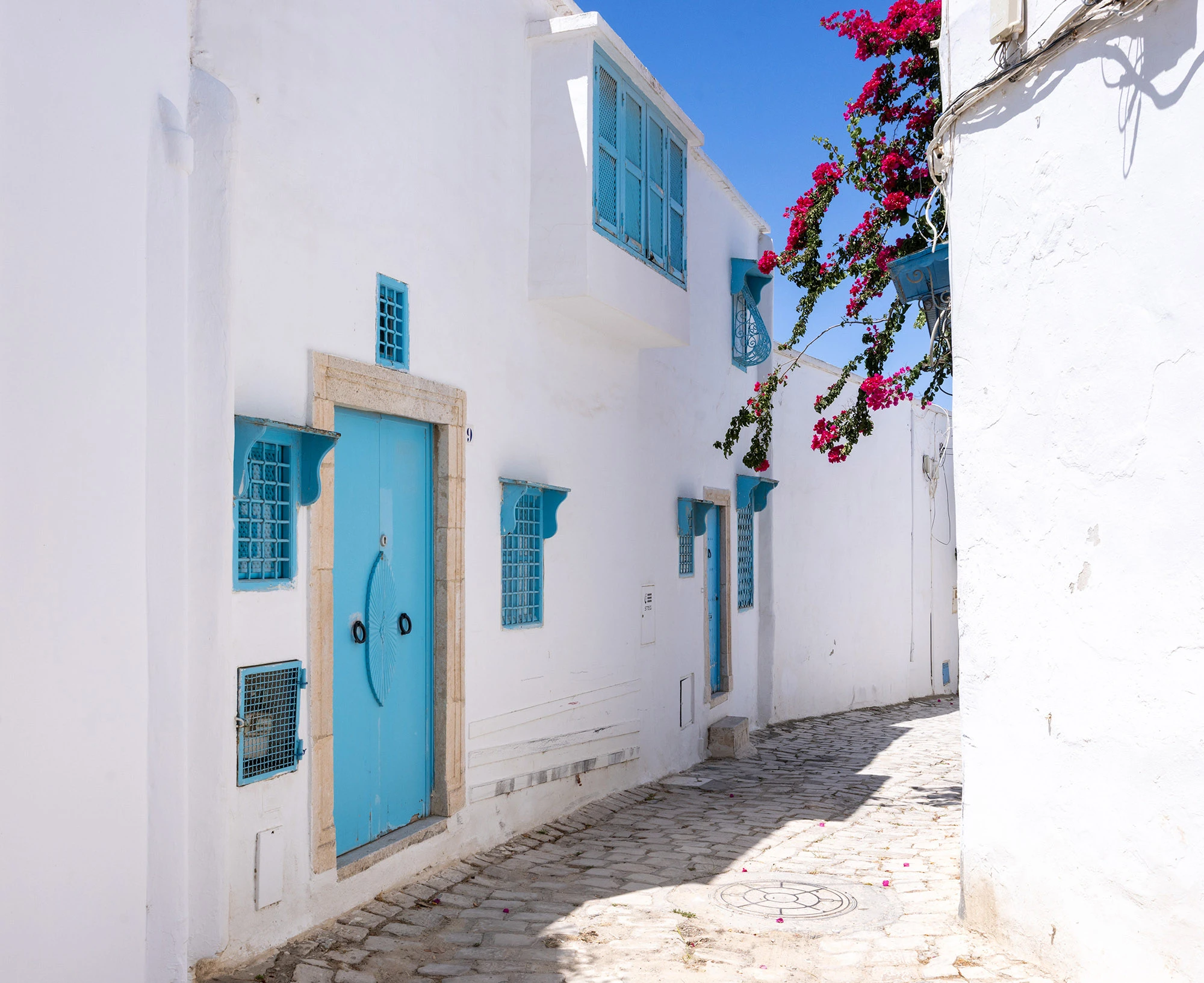 Sunlit alley in Sidi Bou Said, Tunisia, lined with whitewashed walls, turquoise doors, and windows accented by bright pink bougainvillea. The narrow cobblestone path captures the village's harmony of light, color, and scale that makes it feel both real and dreamlike.