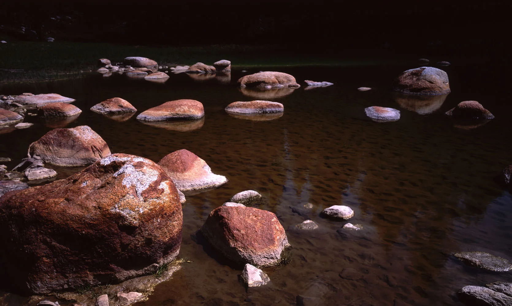Rounded granite boulders resting in the still, tea-dark shallows of a Sierra river, Tuolumne Meadows