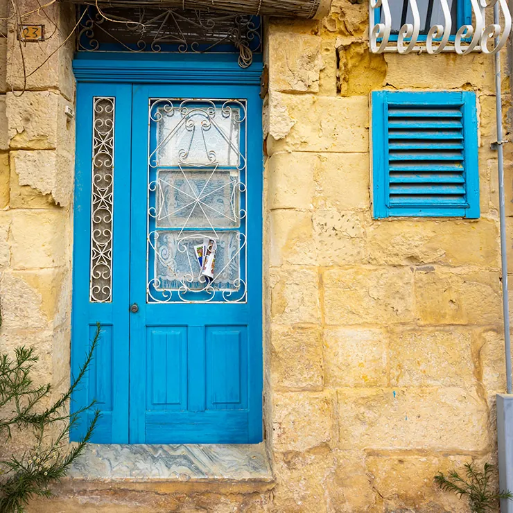 Sky blue wooden door in Valletta, Malta, set in a warm limestone facade, with ornate white wrought iron grilles over glass panels, a small vertical decorative inset, and a matching blue shuttered window nearby.