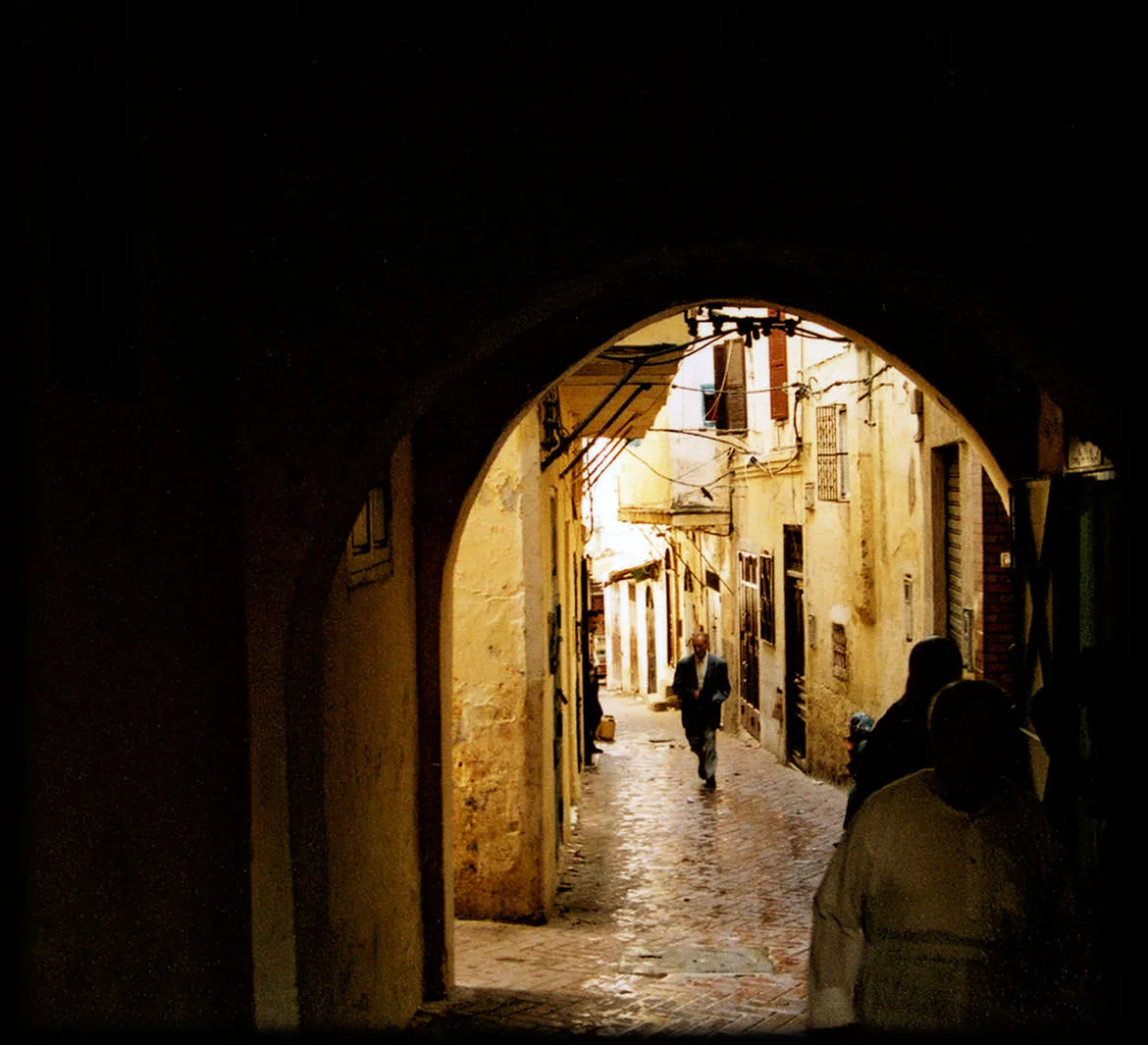 A view through a shadowed stone archway into a sunlit medina alley, a lone figure walking, Tangier