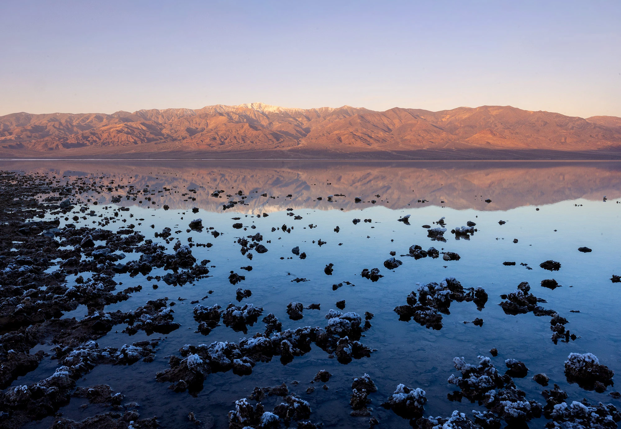 Still water at Lake Manly reflects the Panamint Range and Telescope Peak in Death Valley, with scattered salt formations emerging from the shallow flooded playa.