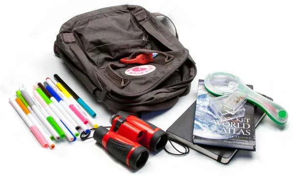 A child's brown Kanken backpack with colored markers, red binoculars, a Moleskine, a pocket world atlas and a magnifier