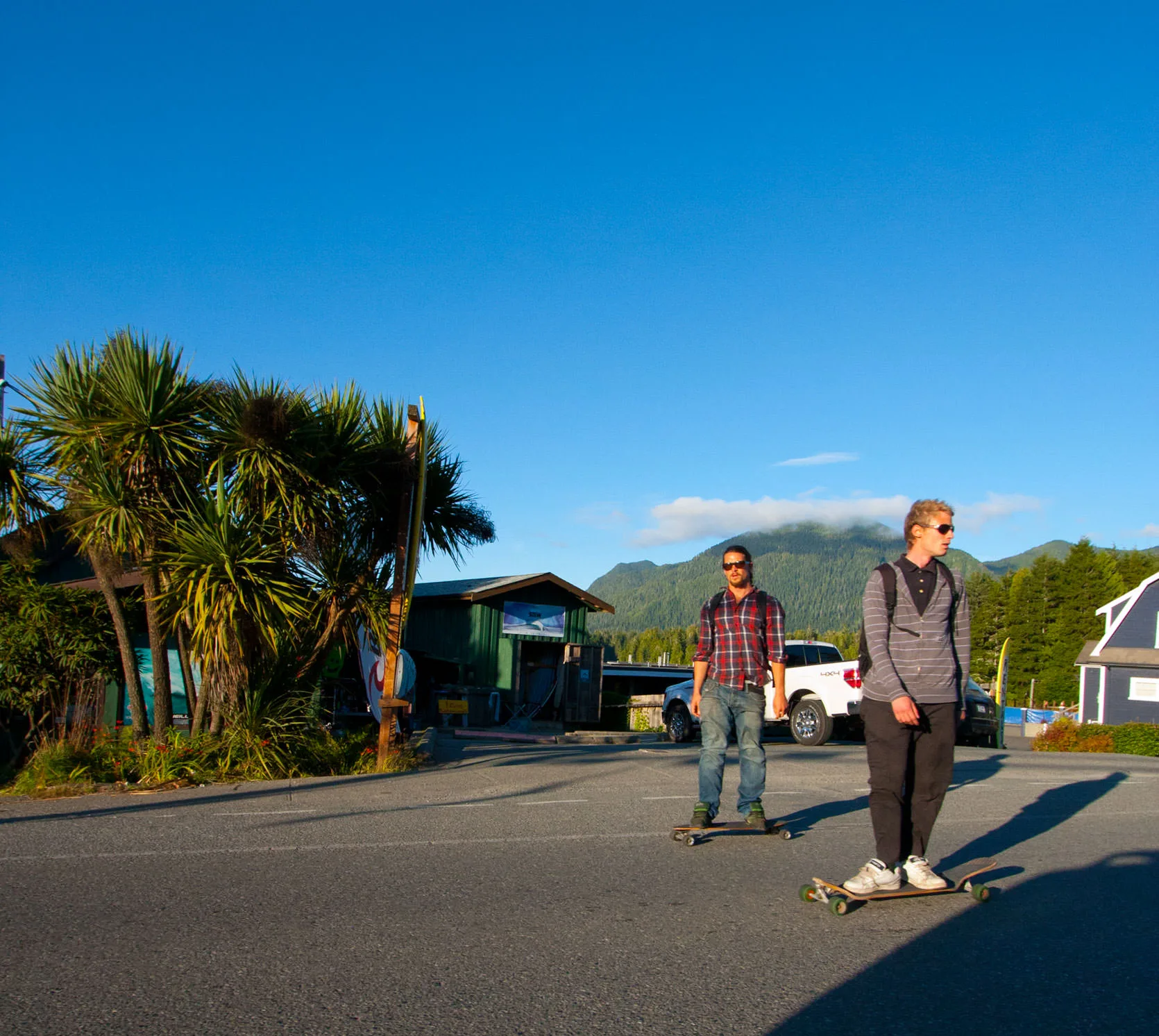 Two young men skateboarding down a sunny street in downtown Tofino, palm-like trees and green mountains behind them.