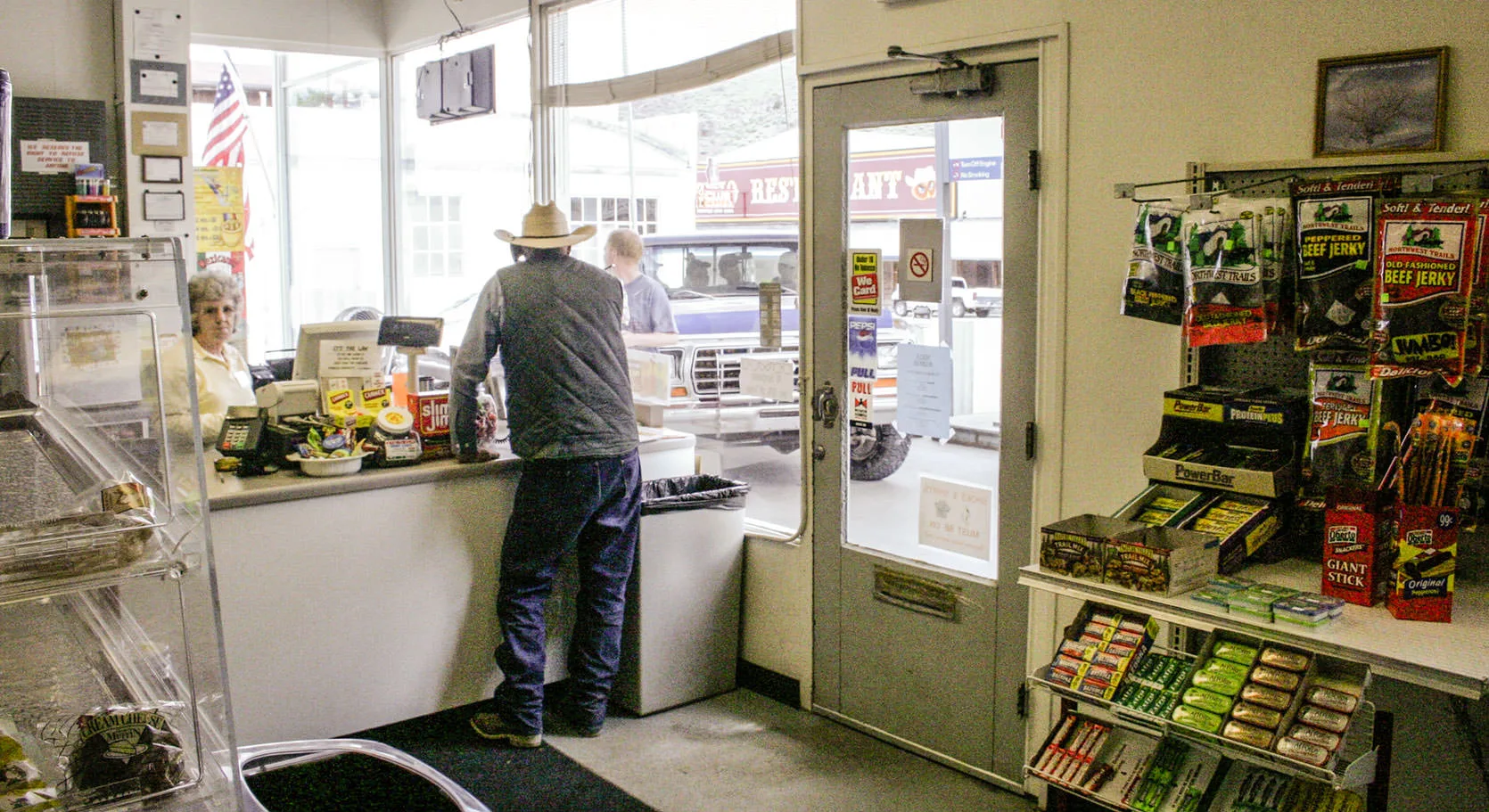 Inside a small-town Nevada market on Highway 50, a rancher in a cowboy hat at the counter beside a rack of beef jerky