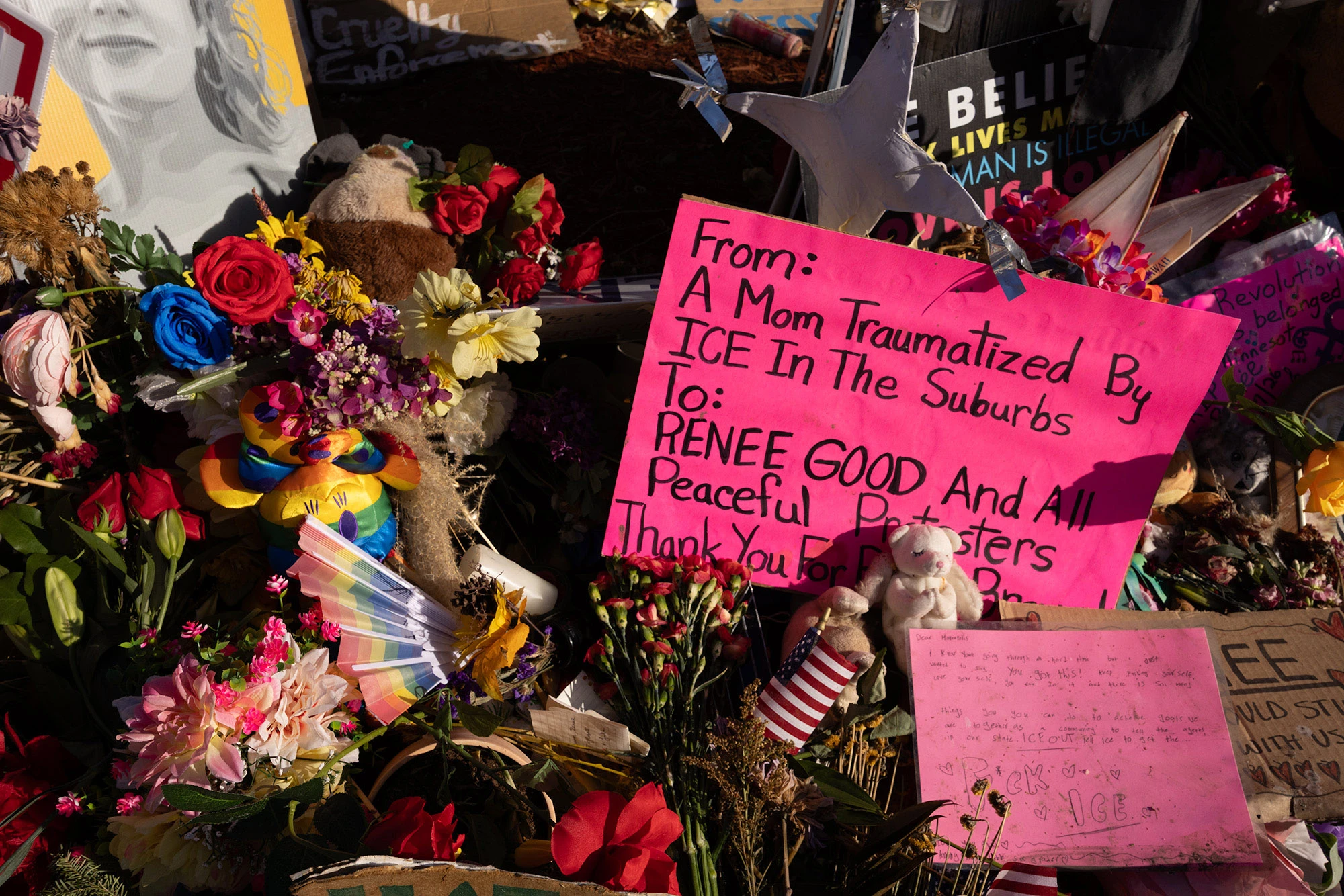 A bright pink protest sign at the Renée Good memorial reads 'From: A Mom Traumatized By ICE in the Suburbs' amid flowers, candles and other tributes.