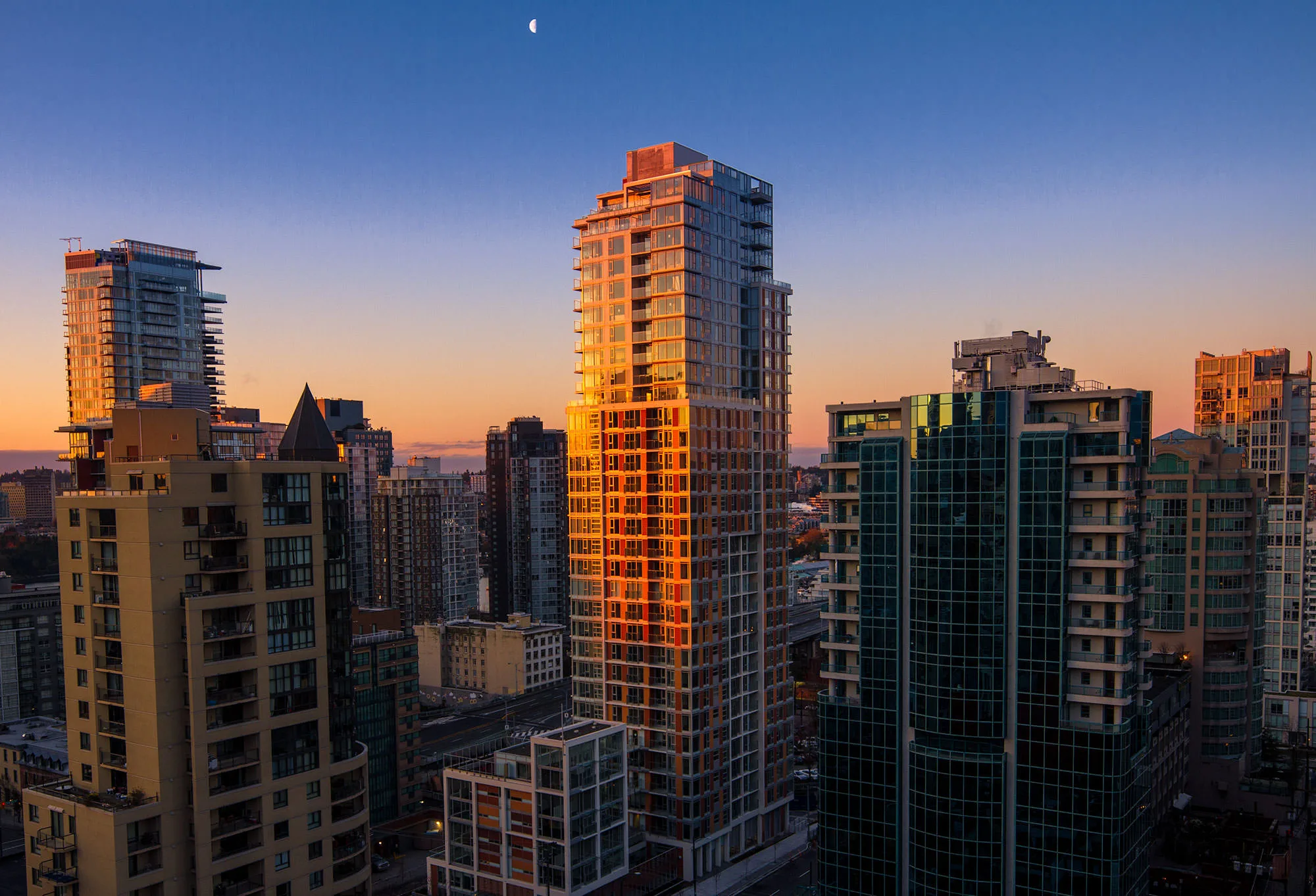 Downtown Vancouver high-rises catching orange evening light against a deep blue sky, the moon above.