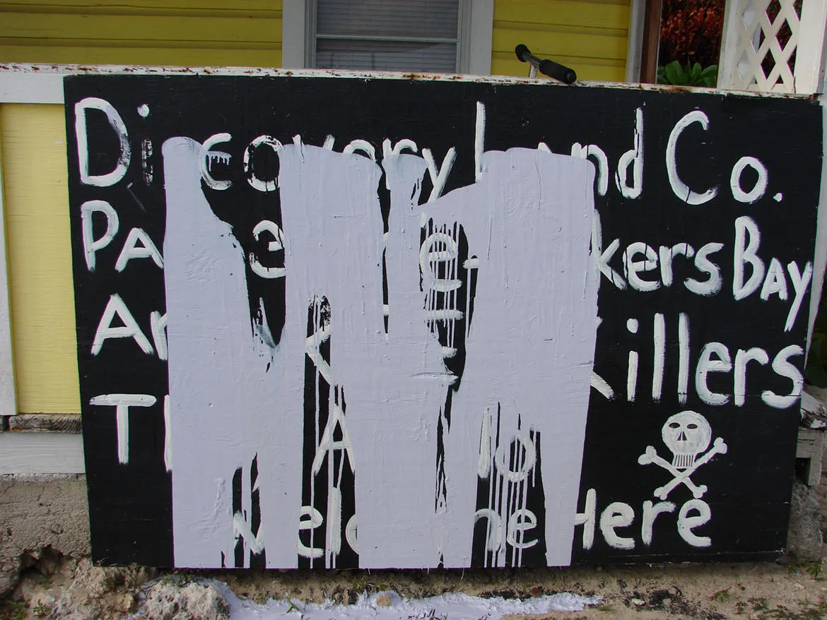 A hand-painted protest sign, partly whitewashed, accusing Discovery Land Co. over Baker's Bay, with a skull and crossbones