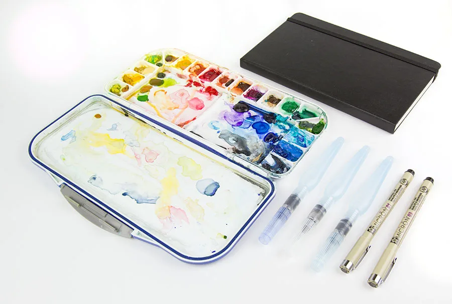 A well-used metal watercolor palette open beside water brushes, Micron pens and a black Moleskine sketchbook