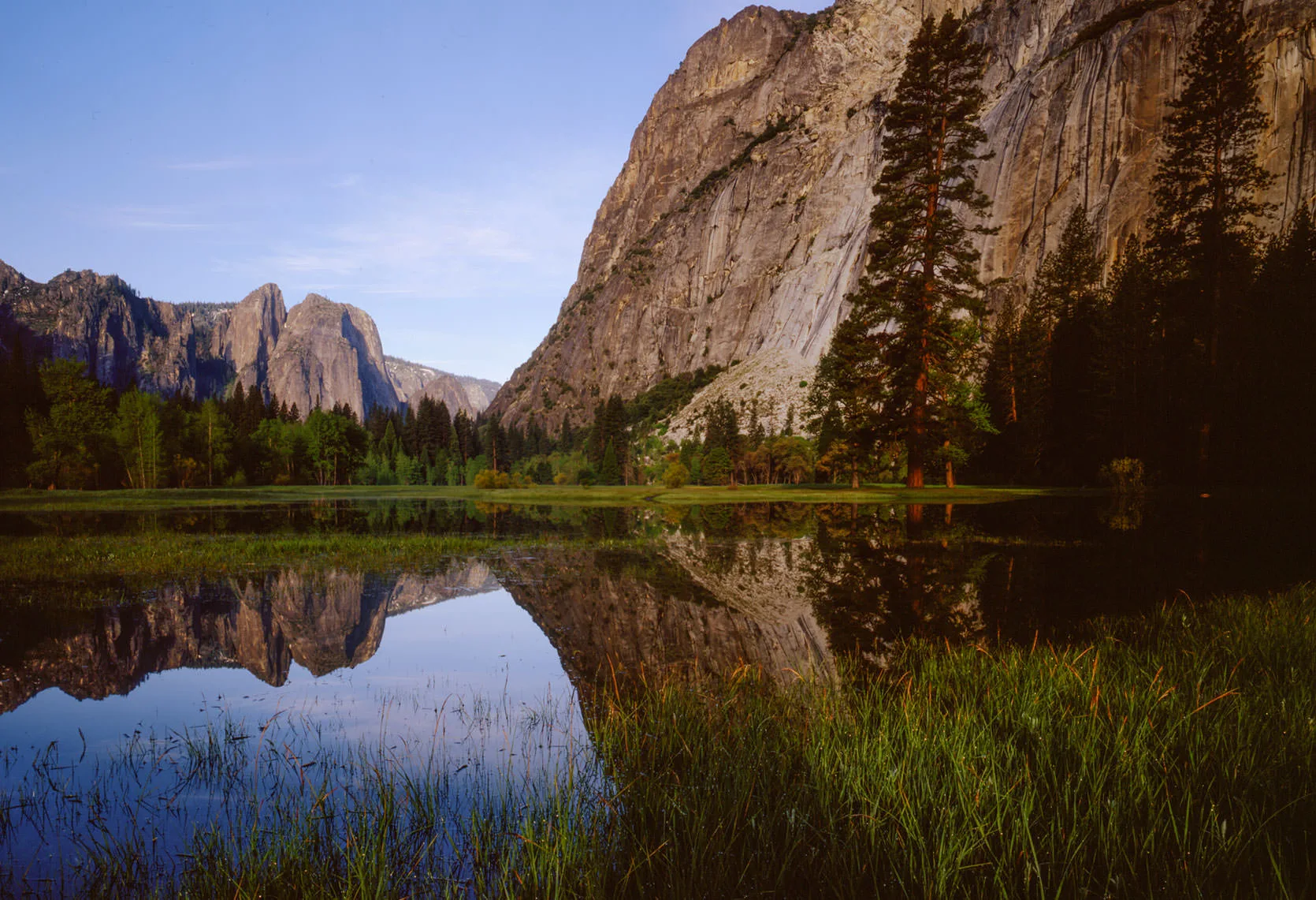 Granite cliffs and a lone tall pine reflected in a calm snowmelt pond in Yosemite Valley, distant Sierra peaks under a clear sky