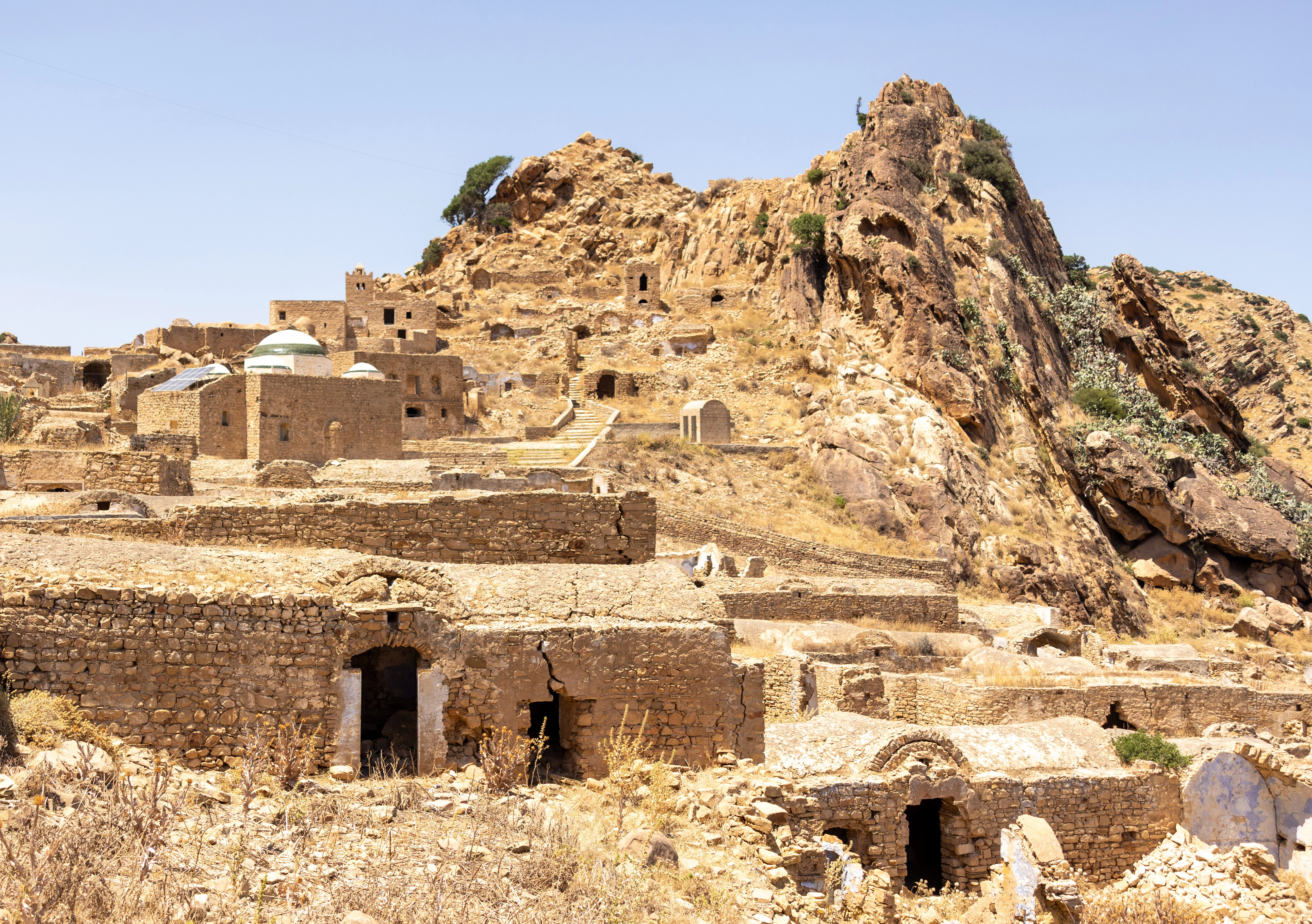 Ancient stone buildings of the abandoned Berber hill village of Zriba Olia in Tunisia, scattered across a rocky mountainside under bright midday light.